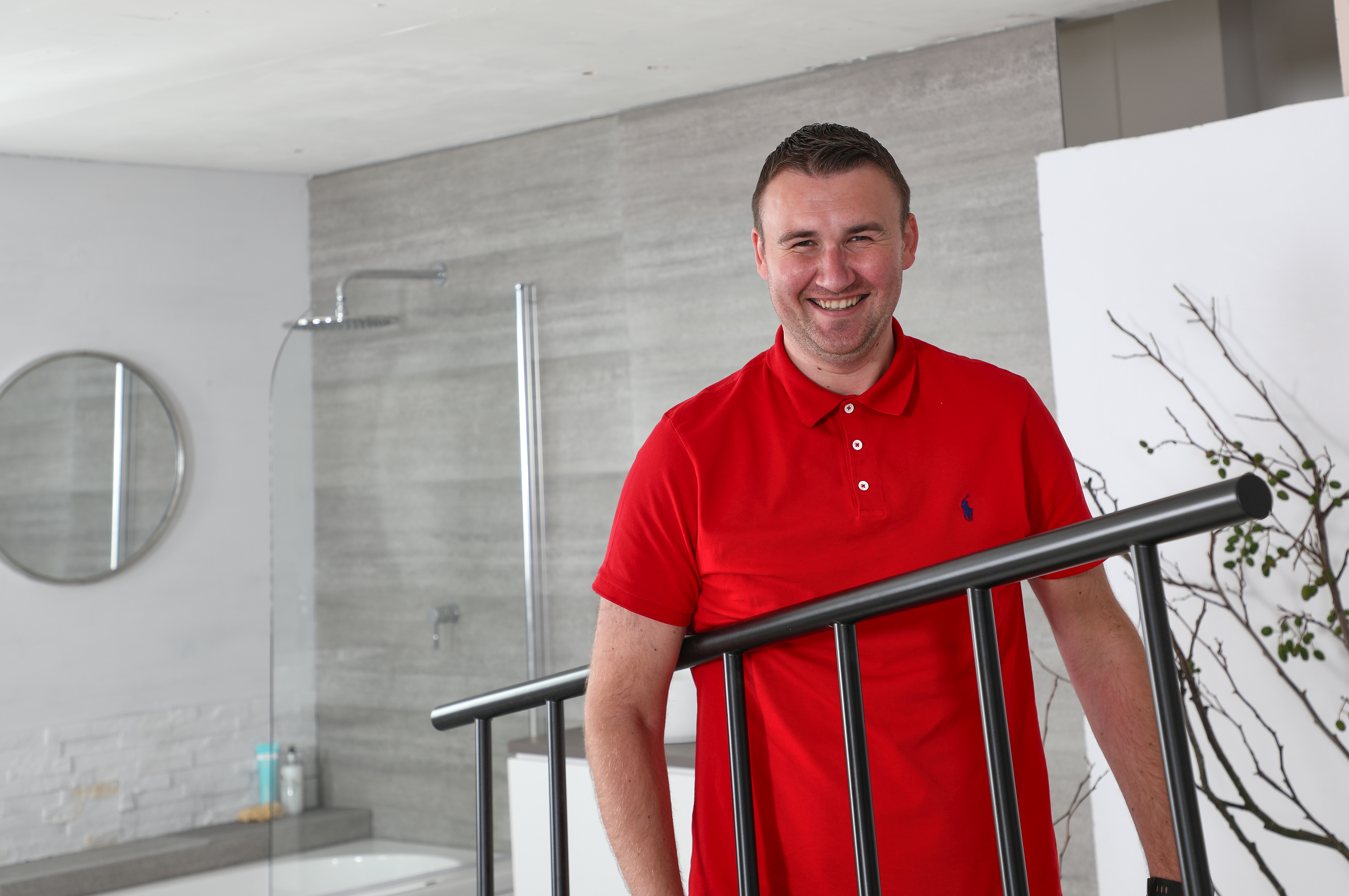 An image of heating technology expert at Bestheating John Klee smiling at the camera wearing a red t-shirt and carrying a black radiator in a bathroom