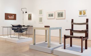 A gallery room with white walls and beige tiled flooring. Multiple framed geometric images on the white wall. In the center of the room are furniture items placed on individual square podiums. A brown chair with horizontal lined textile, A white table with brown square blocks placed on it. A chair with metal frame and black textile and a small glass table with metal base. In the left corner of the room is a black hanging lamp next to a black table with silver legs. Placed on the table are 1 black plate and a black bowl
