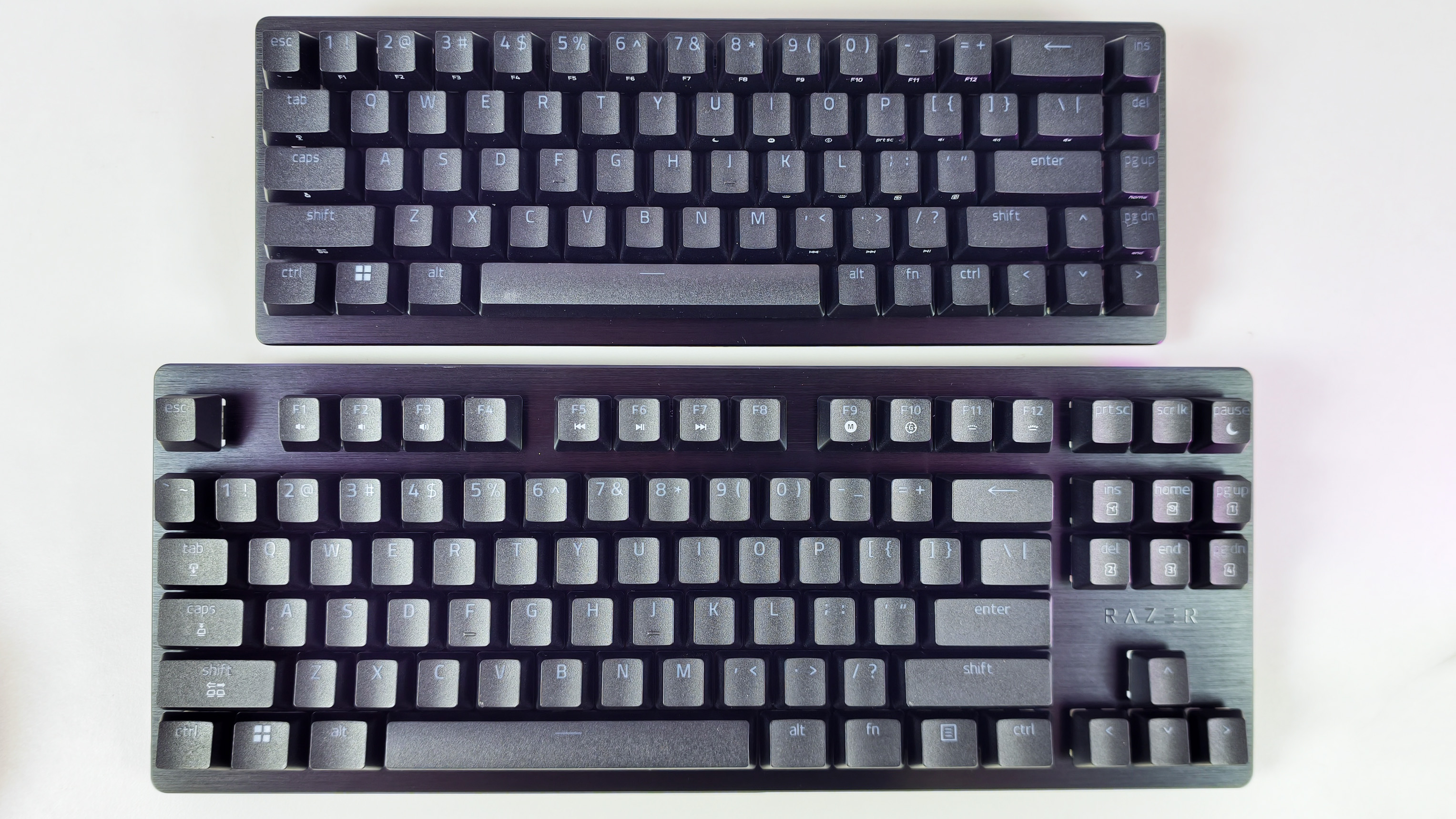 Top-down view comparing the compact Razer Huntsman V3 HE Mini 65% keyboard (top) and the larger TKL keyboard (bottom) on a white desk.