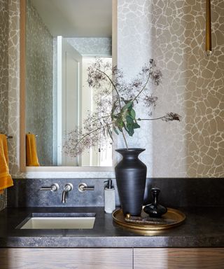 Small bathroom with stone wallpaper, dark countertop, vase, orange towel, rectangular mirror,