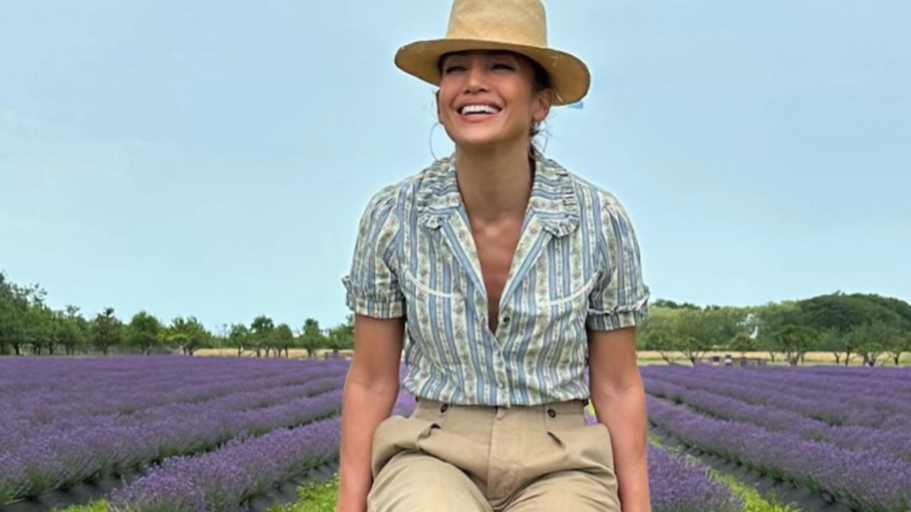 Jennifer Lopez sits in front of a lavender field wearing a Ralph Lauren floral top and straw hat, complementing her Ralph Lauren pink maxi dress and wedge sandals