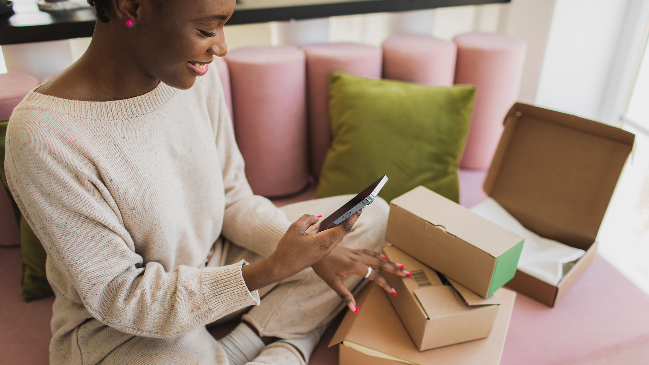 A smiling woman holding a smartphone whilst sitting on a pink sofa with cardboard boxes