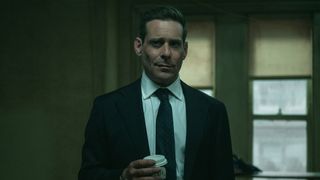 James Callis wears a suit and holds a cup of coffee while standing in front of two windows in Slow Horses season 5 episode 6