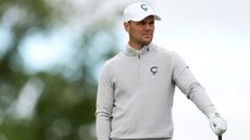 Martin Kaymer resting his hand on his club while wearing a grey pullover and white cap