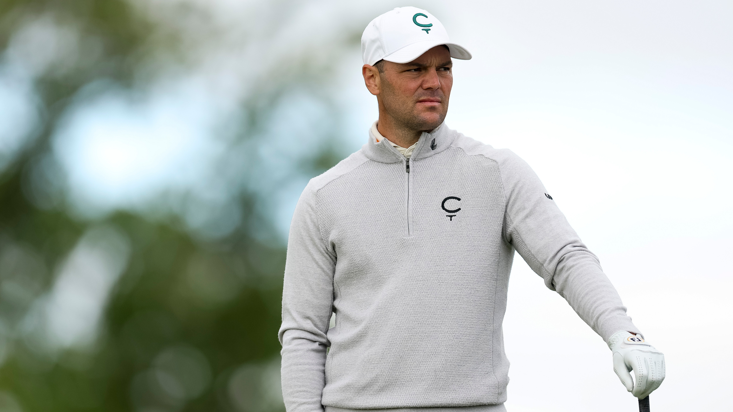 Martin Kaymer resting his hand on his club while wearing a grey pullover and white cap