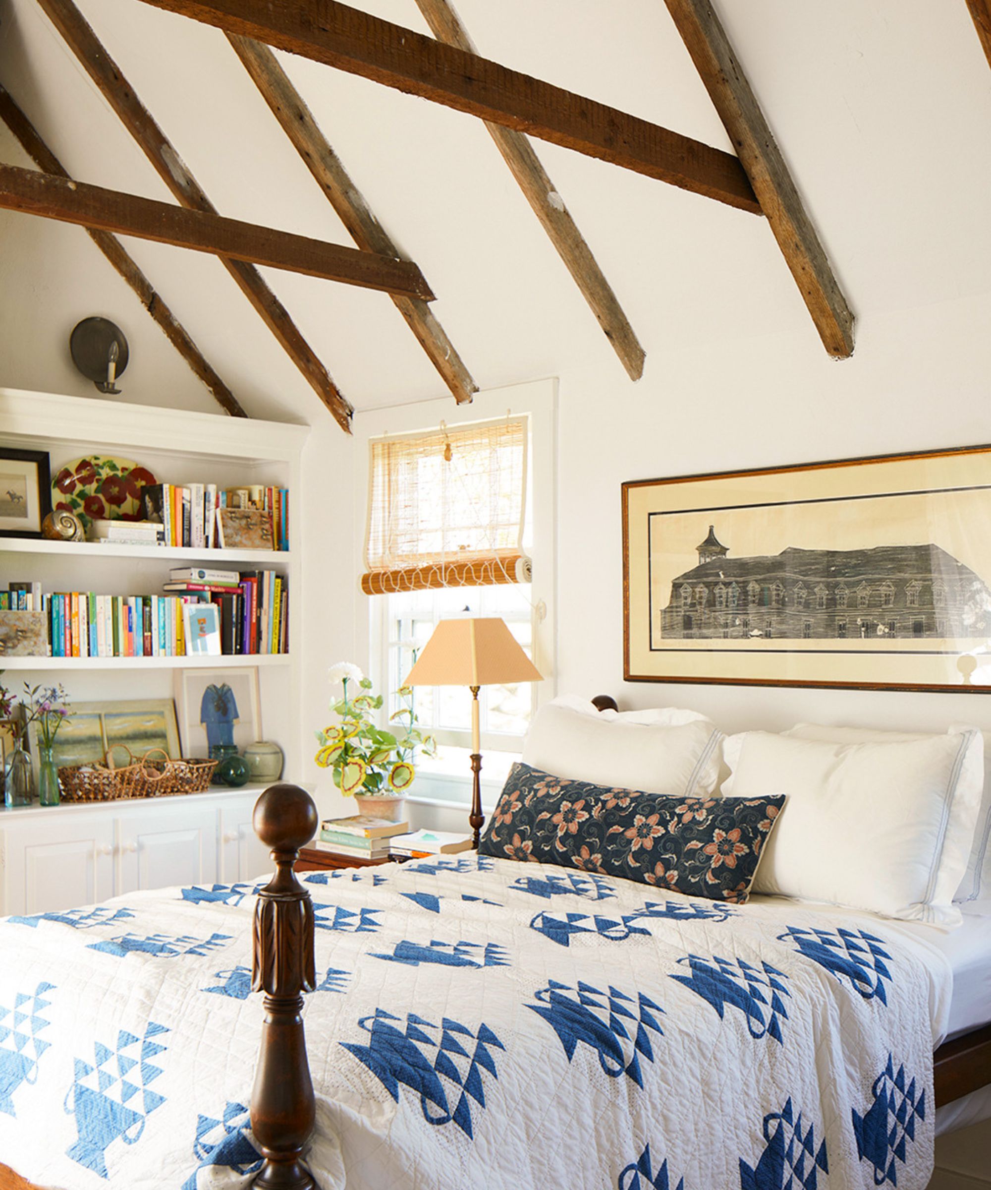 Bright bedroom with exposed wooden beams, built-in white shelving full of books, and a bed covered with a white and blue geometric quilt