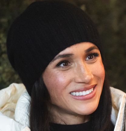 Meghan Markle wearing a black winter hat and a white puffer coat smiling and looking to the right