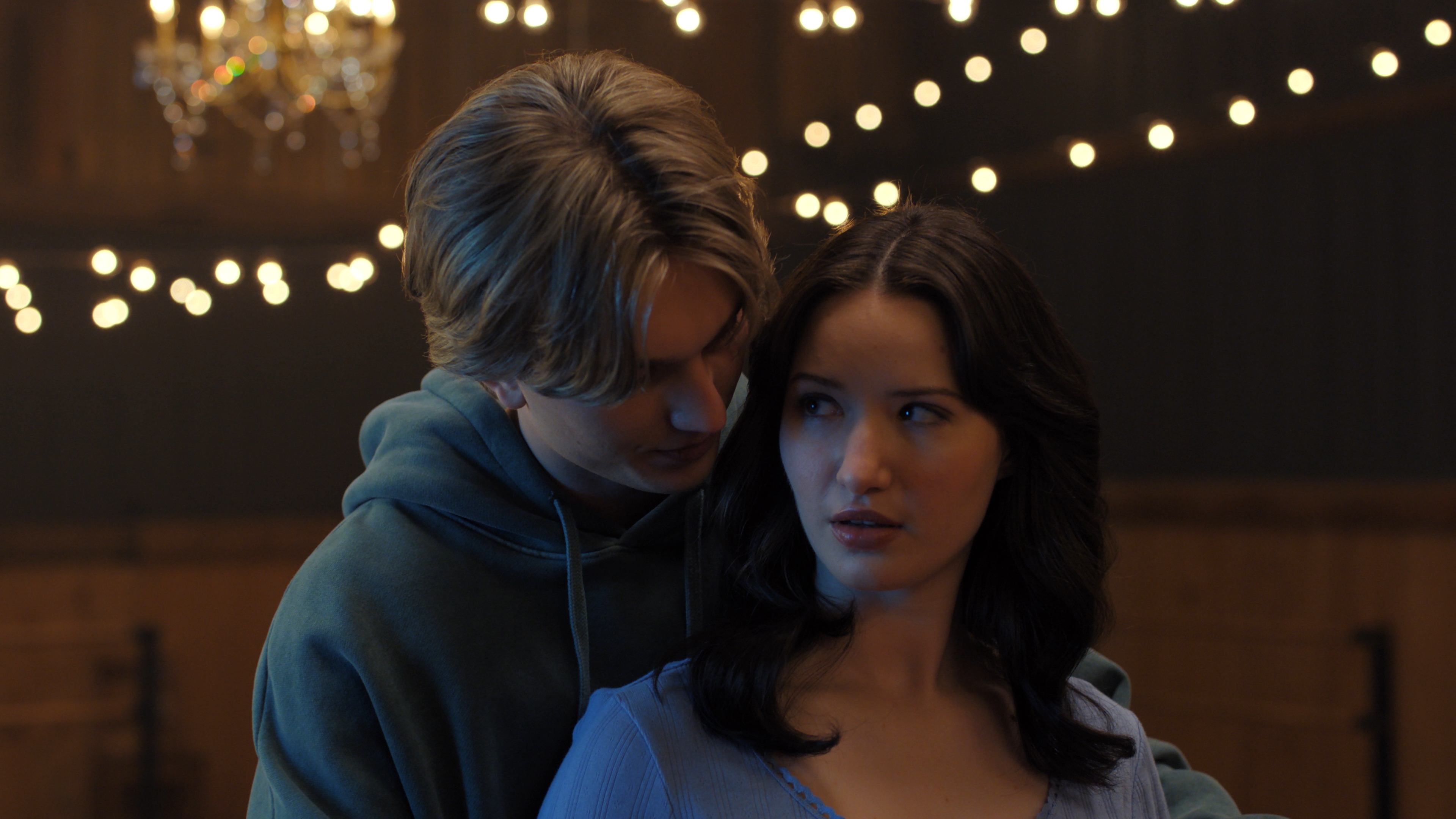 Cale Ambrozic as Brayden Elliot wearing a sweatshirt and embracing Madelyn Keys as Adriana Russo from behind while on an ice rink lit in twinkly lights in Finding Her Edge: Season 1
