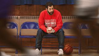 Adam Sandler sidder på en stolerække med en kaffe i hånden og en basketbold ved den ene fod fra filmen Hustle