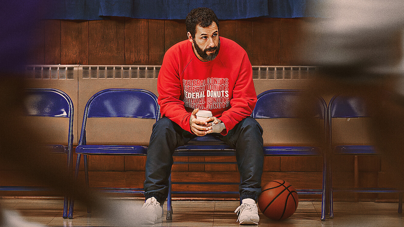 Adam Sandler sidder på en stolerække med en kaffe i hånden og en basketbold ved den ene fod fra filmen Hustle