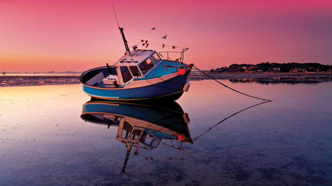 A small sailing boat with a mirrored reflection in the water, shot in low light conditions at dusk. The sky is a vivid pink and provides a good contrast against the blue water