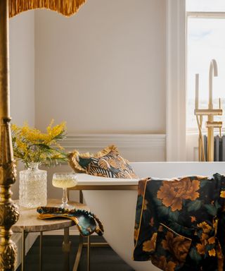 A bathoom with a gold fringed lamp, a marble side table with a glass bottle and cocktail glass, and a bath tub with a dark blue, white and gold throw pillow in it and a matching blanket hanging from the edge