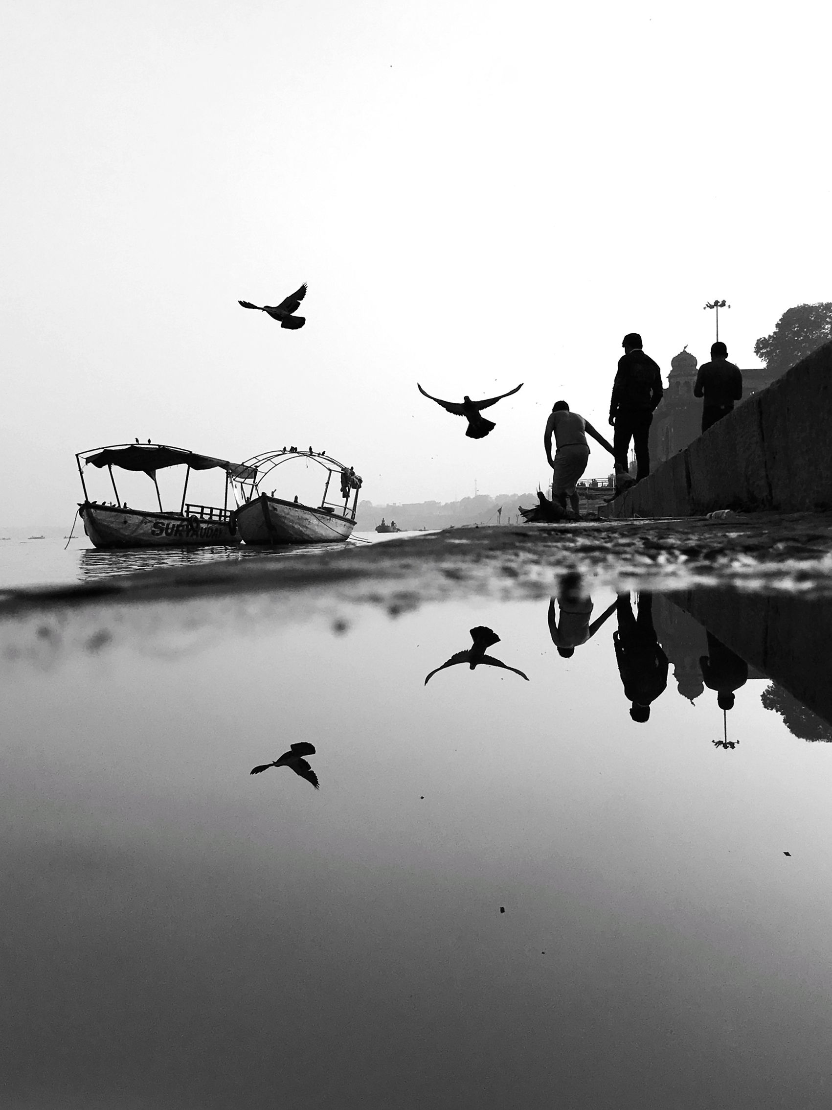 Black-and-white image of a riverside scene with boats, two people walking, and birds flying overhead. Their reflections are visible in a water puddle