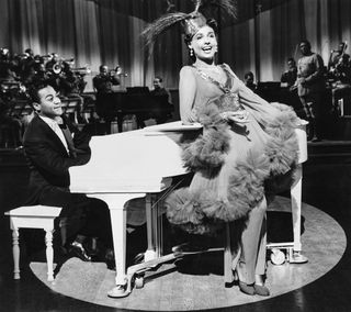 a woman in a feather dress sings while leaning against a piano on stage in Stormy weather