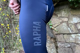 The Rapha Pro training tights reflective RAPHA logo can be seen close up on the leg of a rider who is standing in front of a stone wall with greenery overhanging.