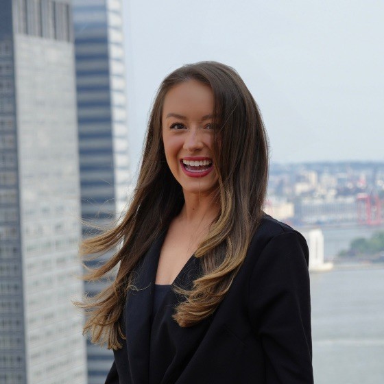 Photograph of Hannah Pregont smiling in black smart attire in front of blurred-out city background.