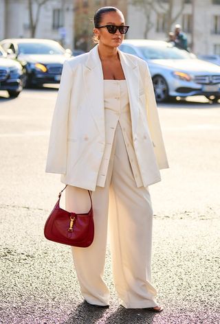 a street style photo of a woman wearing one of the most popular vintage bags, Gucci Jackie shoulder bag in red styled with an all-white suit, white sandals, and black sunglasses