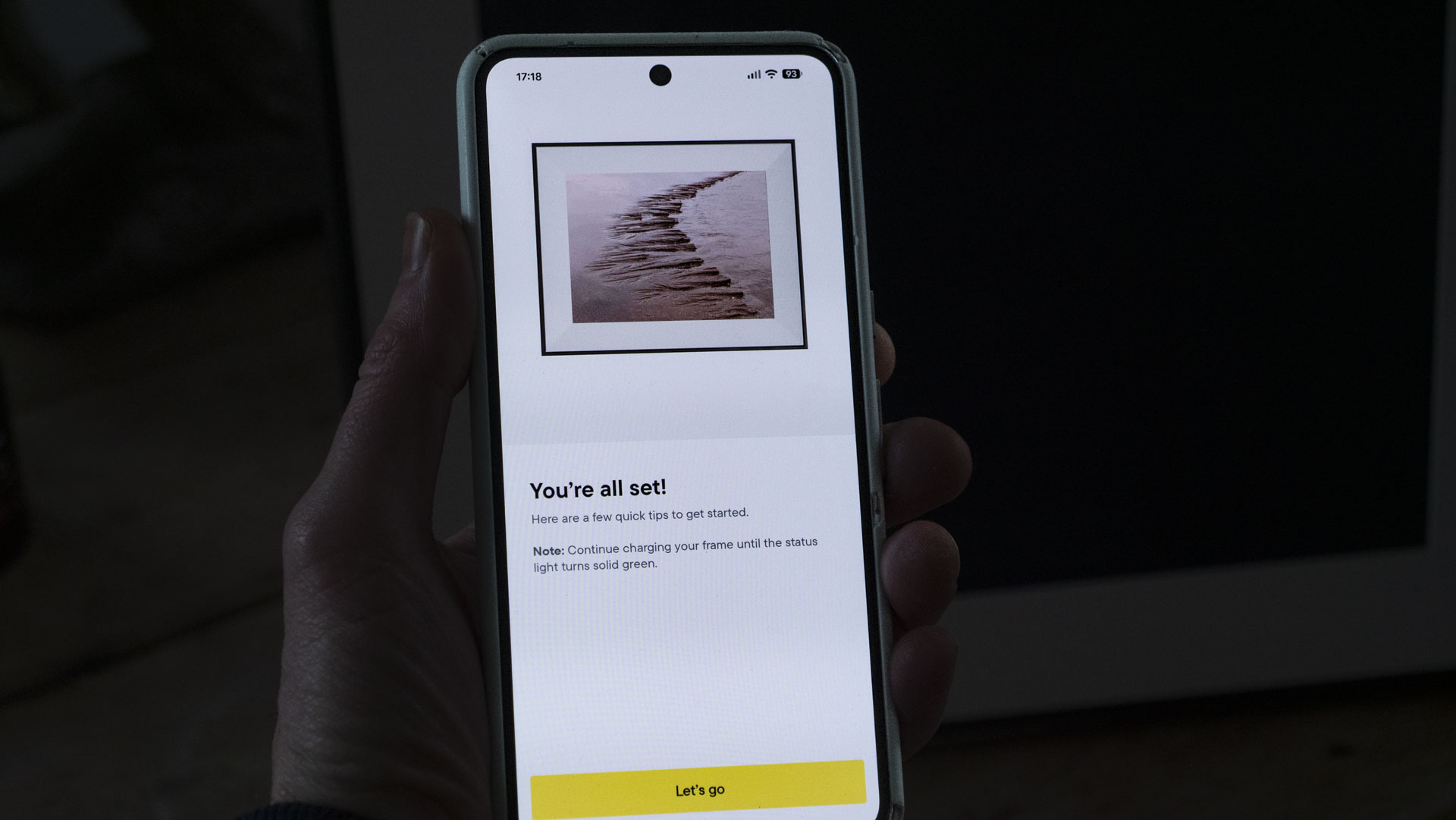 A phone being held, displaying the Aura companion app for digital photo frames, in front of the The Aura Ink 'E Ink' digital photo frame on a sideboard
