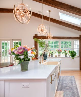 A white kitchen island with colorful plants on it, a gold tap, with a window ledge with colorful plants beyond it and three gold circular hanging pendant lights on the white ceiling with dark wooden beams