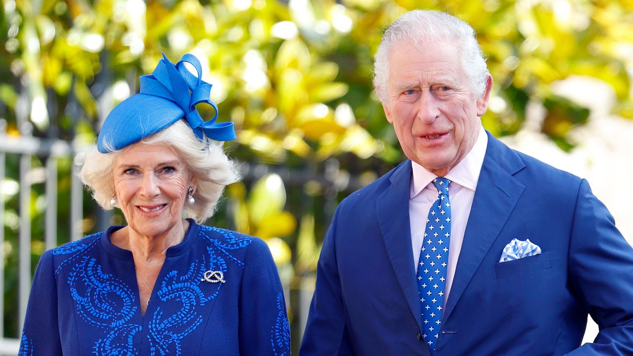 Queen Camilla wearing a royal blue dress and matching hat smiling with King Charles who is wearing a blue suit and a blue and white polka dot tie