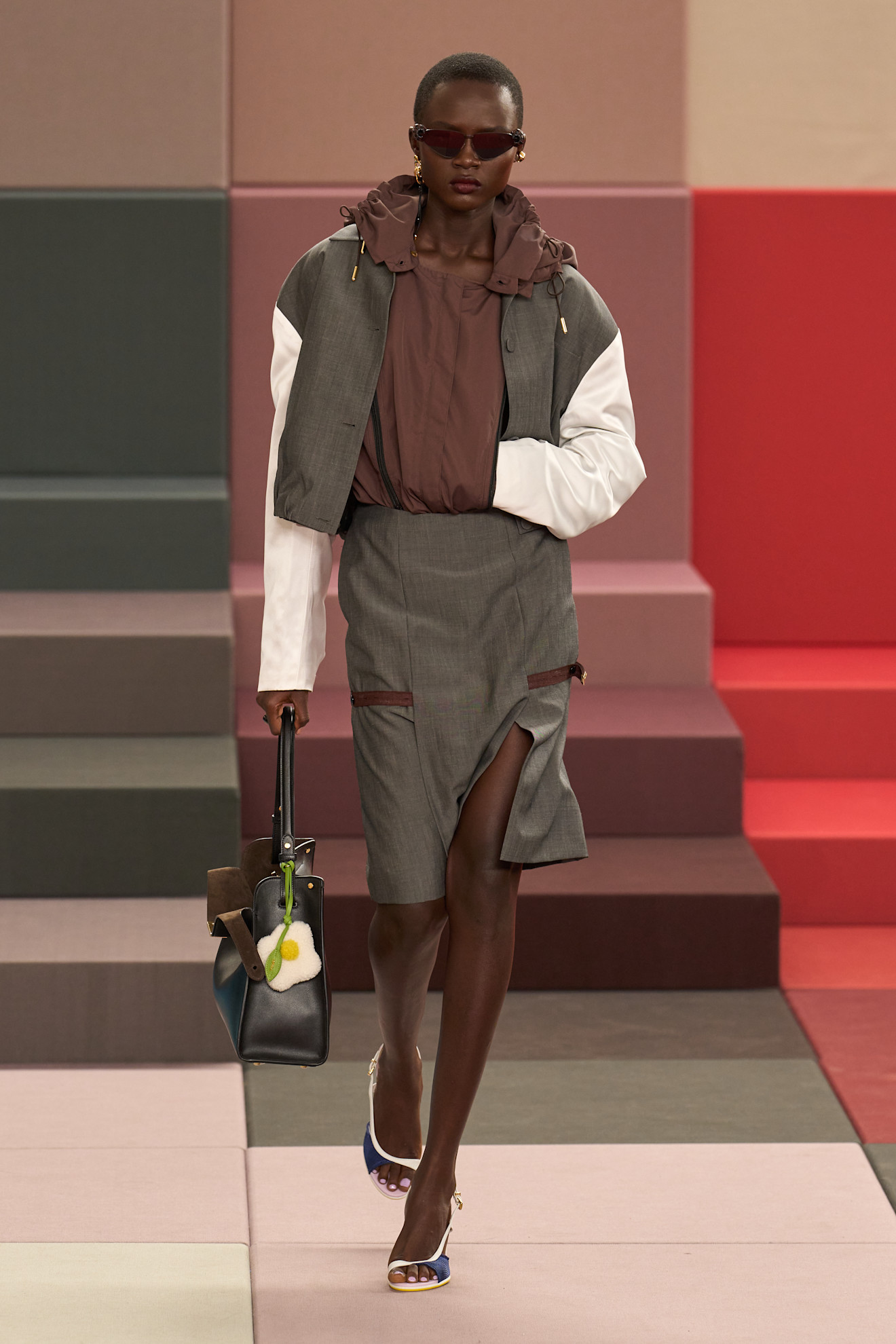 Layering staples for spring 2026 are shown in a photo from Fendi's runway show, with a model walking while wearing a gray bomber over a dark brown parka jacket that's tucked into a gray pencil skirt styled with two-tone sandals, brown sunglasses, and a black handbag