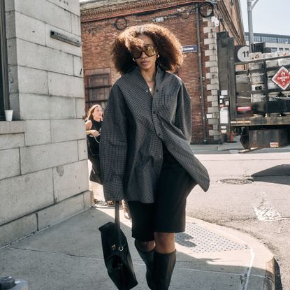 Chic woman street style wearing oversized sunglasses with an oversized button down top, black knee-length shorts and knee high boots with a black bag 