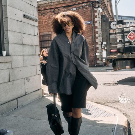 Chic woman street style wearing oversized sunglasses with an oversized button down top, black knee-length shorts and knee high boots with a black bag 