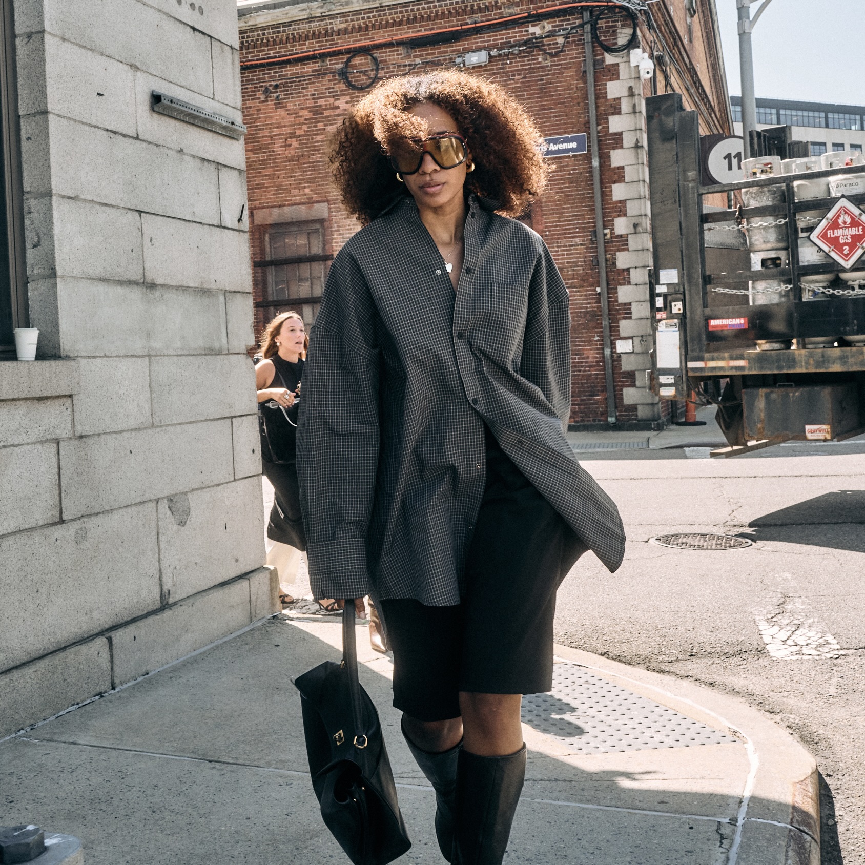 Chic woman street style wearing oversized sunglasses with an oversized button down top, black knee-length shorts and knee high boots with a black bag 