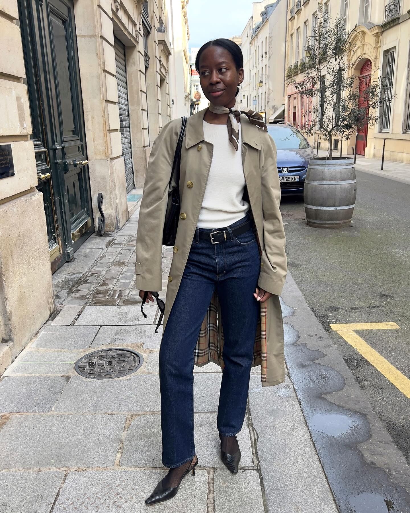 Style influencer Sylvie Mus poses in Paris wearing a printed silk neck tie scarf, a basic white T-shirt, a trench coat, dark-wash straight jeans, and black slingback heels.