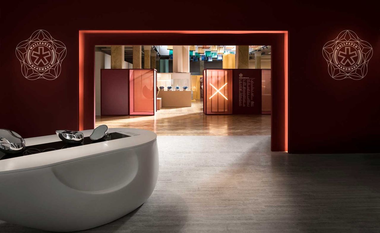 Installation view of Wallpaper* Handmade X featuring wine red walls with Wallpaper* Handmade branding, a white curved structure with silver pieces on top and an illuminated &#039;X&#039; in the distance