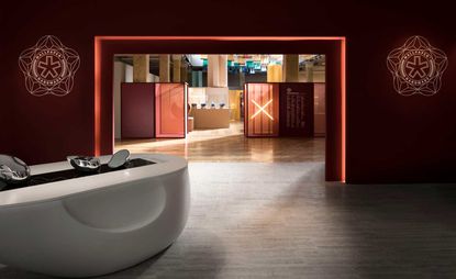 Installation view of Wallpaper* Handmade X featuring wine red walls with Wallpaper* Handmade branding, a white curved structure with silver pieces on top and an illuminated 'X' in the distance