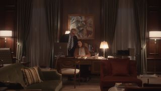 Joan Allen as Sheila Mullen sitting at her desk looking at a computer with husband George standing behind her in a sepia toned, expensively furnished room at their home.