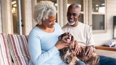 An older couple smile while they sit on the porch and interact with their dog.