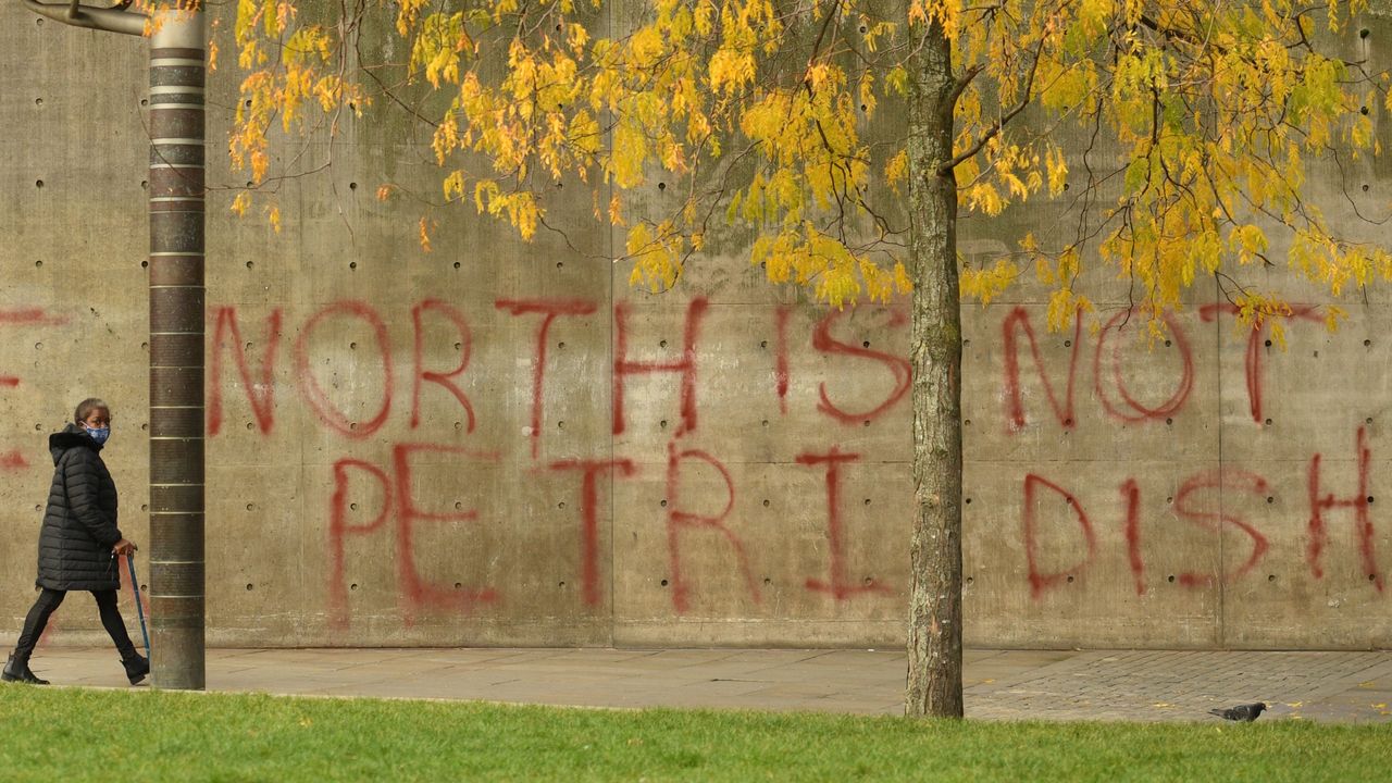 A pedestrian wearing a face-mask walks past graffiti declaring that &amp;#039;the north is not a petri dish&amp;#039;.