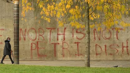 A pedestrian wearing a face-mask walks past graffiti declaring that 'the north is not a petri dish'.