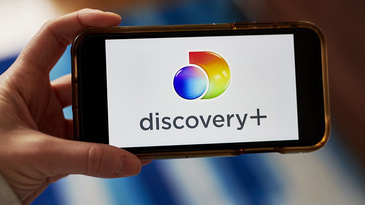 A left hand holding a smartphone with the Discovery Plus logo on it