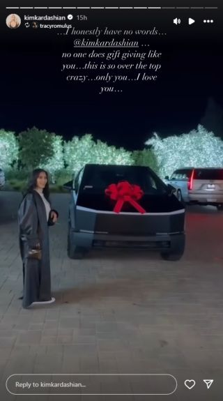 Kim Kardashian smiling with a black Cybertruck with a red bow on it behind her.