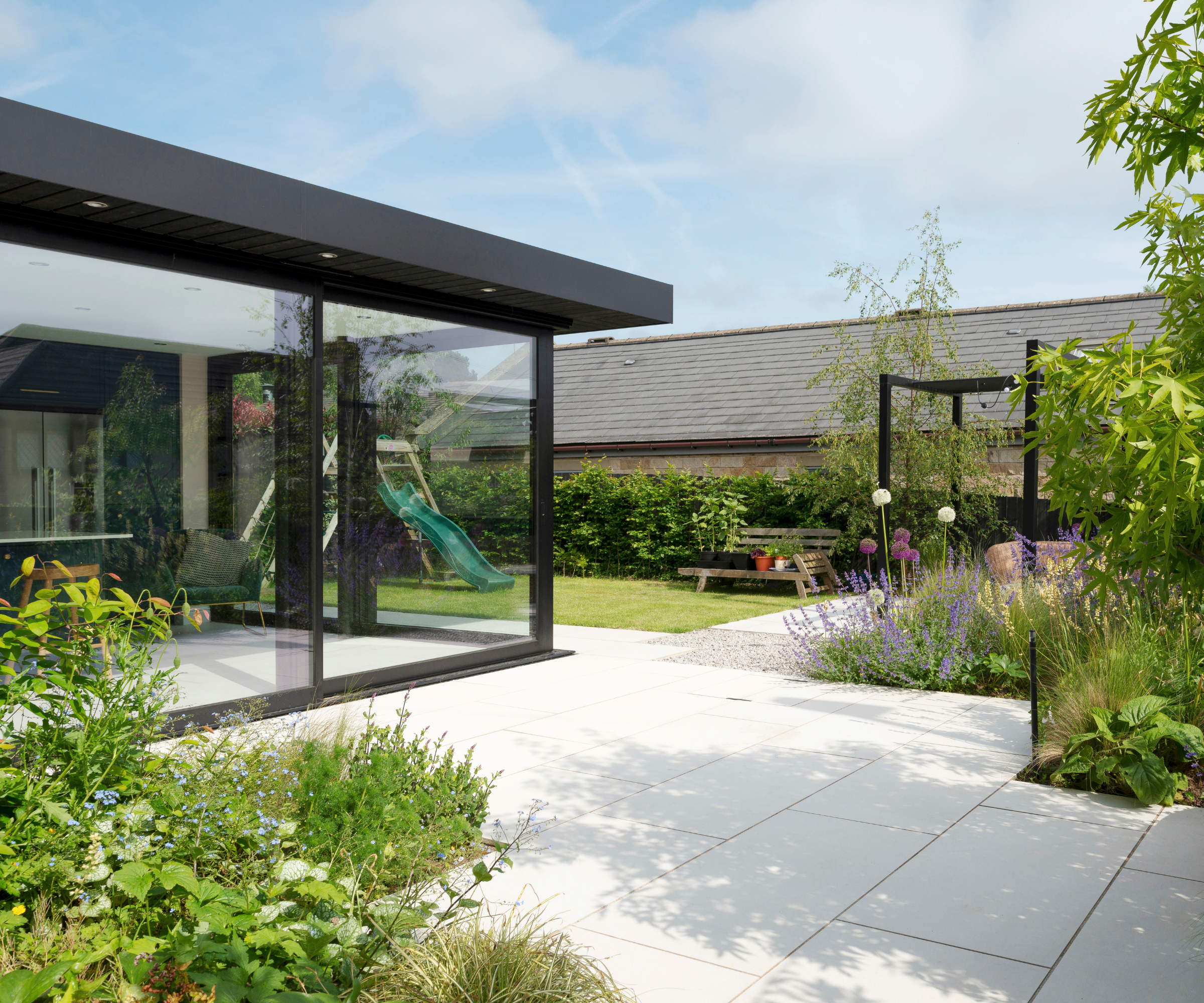 Back garden with glass box extension, patio, lawn with a children's slide, and flower beds