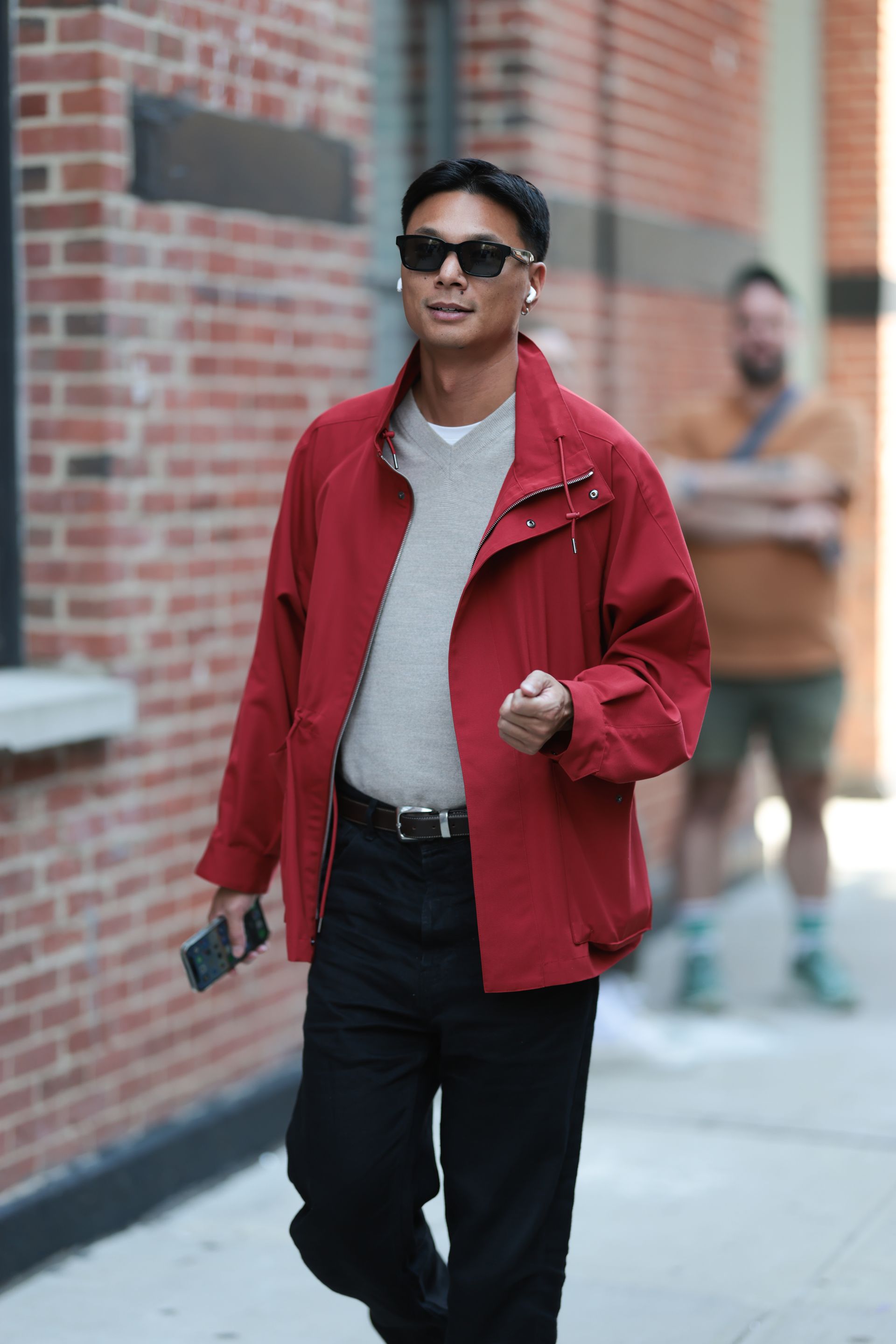A guest seen wearing black rectangular sunglasses, white wireless Apple Airpods earphones, a bold red zip-up jacket layered over a light gray v-neck wool sweater and white cotton basic shirt, black trousers, and a black leather belt outside the Lafayette 148 show during New York Fashion Week on September 11, 2025 in New York City.