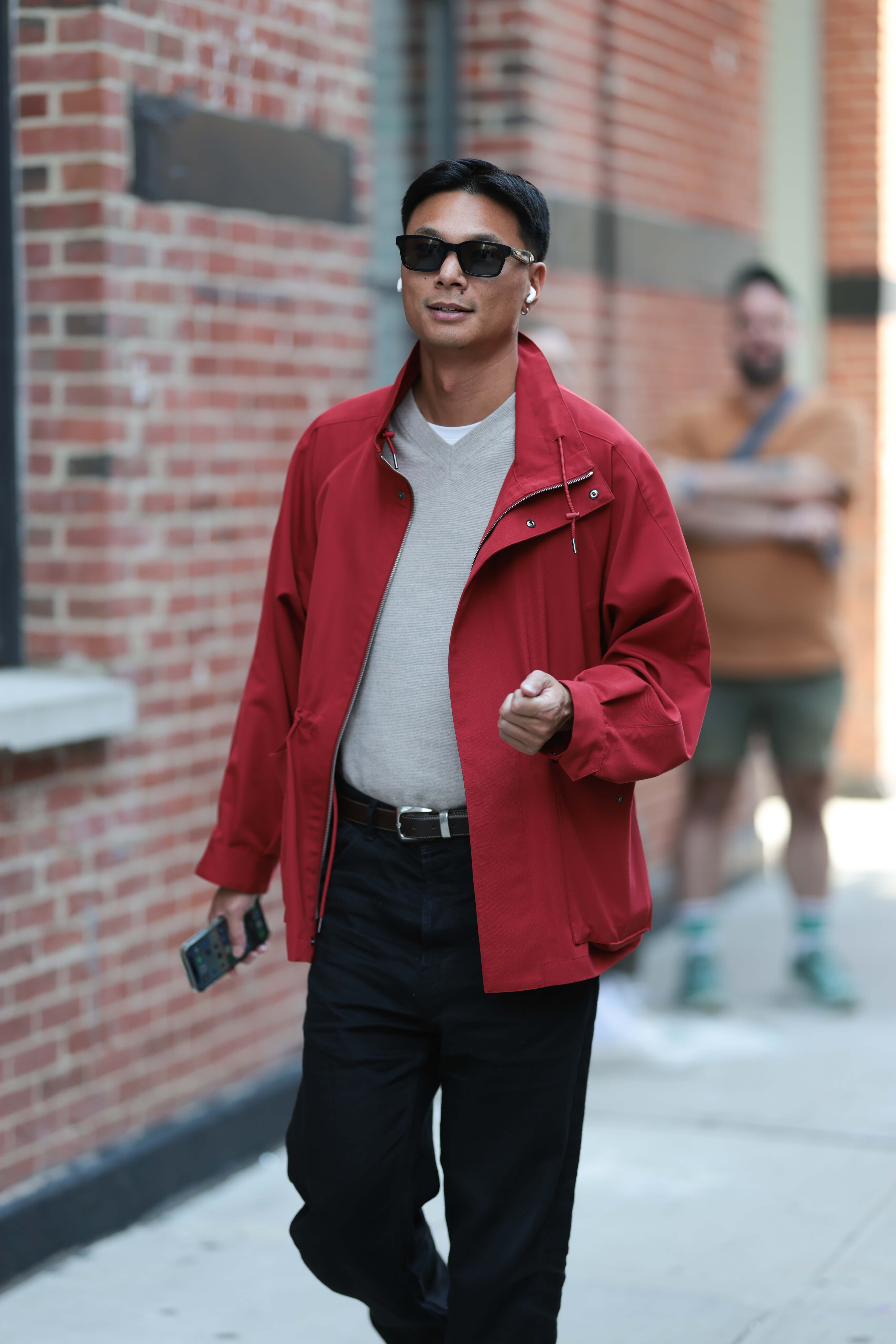 A guest seen wearing black rectangular sunglasses, white wireless Apple Airpods earphones, a bold red zip-up jacket layered over a light gray v-neck wool sweater and white cotton basic shirt, black trousers, and a black leather belt outside the Lafayette 148 show during New York Fashion Week on September 11, 2025 in New York City.