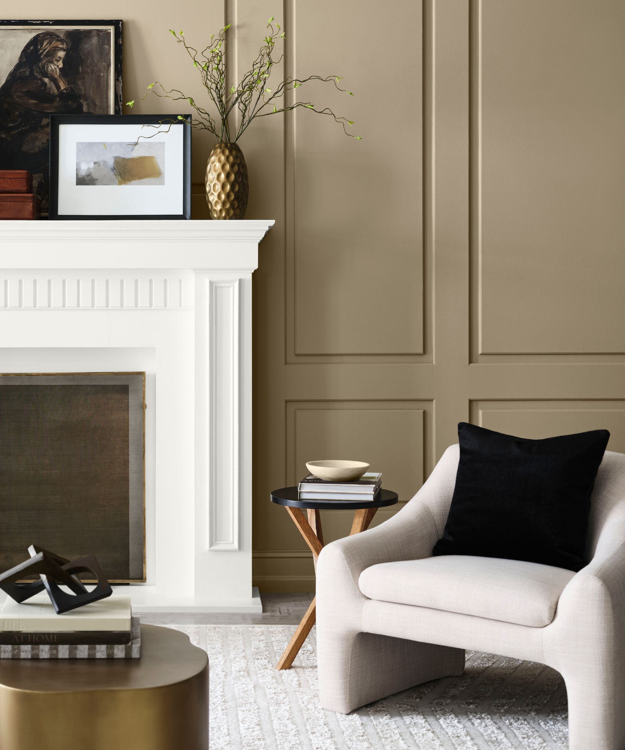 Living room painted in Sherwin-William Universal Khaki with a white fireplace. The mantel is decorated with frames and a there is a cream armchair with a small wood side table