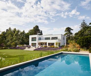 A modern Bauhaus-style self build, clad in white render and sitting within a landscaped plot with a swimming pool. The property, built by Baufritz, features a flat roof with solar panels.