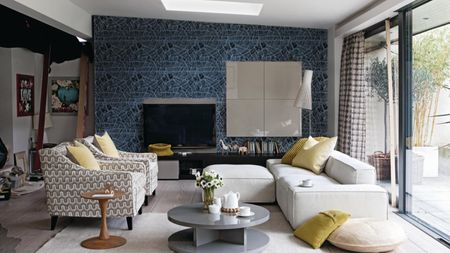 Gray living room, grey graphic patterned wallpaper, white fabric sofa with matching footstool, armchairs upholstered in hexagonal patterned fabric , yellow scatter cushions, grey veneer coffee table, lacquer Boiserie TV panel, lacquer four cube wall unit, sliding patio doors
