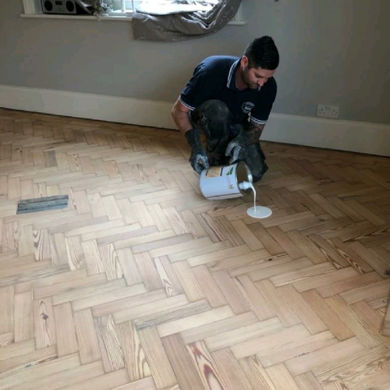 David Clarke, the owner of Absolute Floor Sanding Ltd., is shown pouring a wood finishing product onto a herringbone parquet floor.