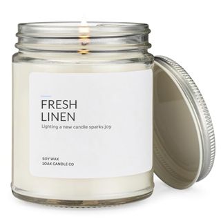 A clear glass jar with a white candle inside and a metal screw lid leaning against it. A white label with 'fresh linen' printed on the front. 