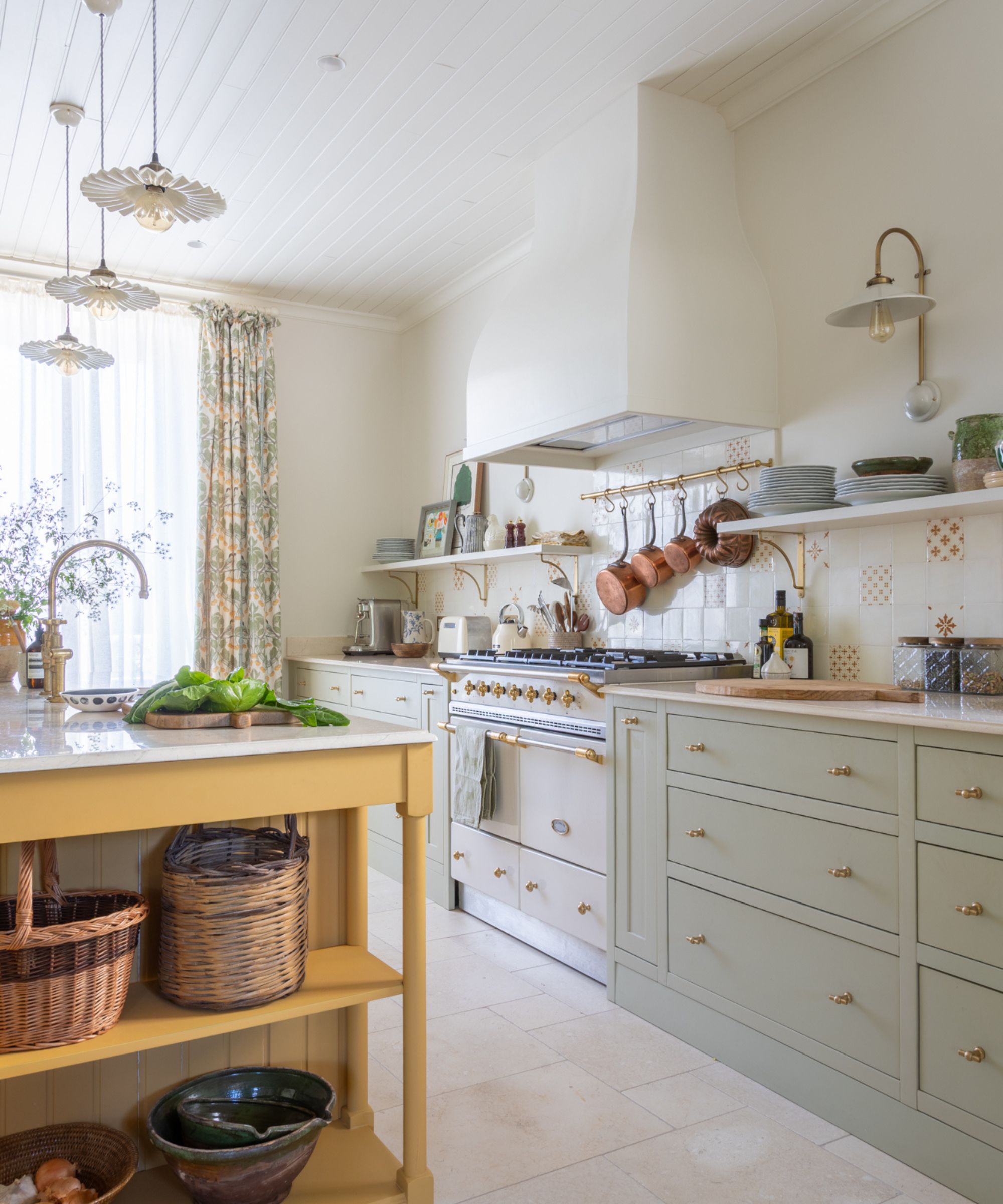 A bright, cottage-style kitchen featuring pale sage green lower cabinets, a cream-colored range cooker, and a mustard yellow central island with woven baskets on open shelving. Copper pots hang above the stove against a patterned tile backsplash.