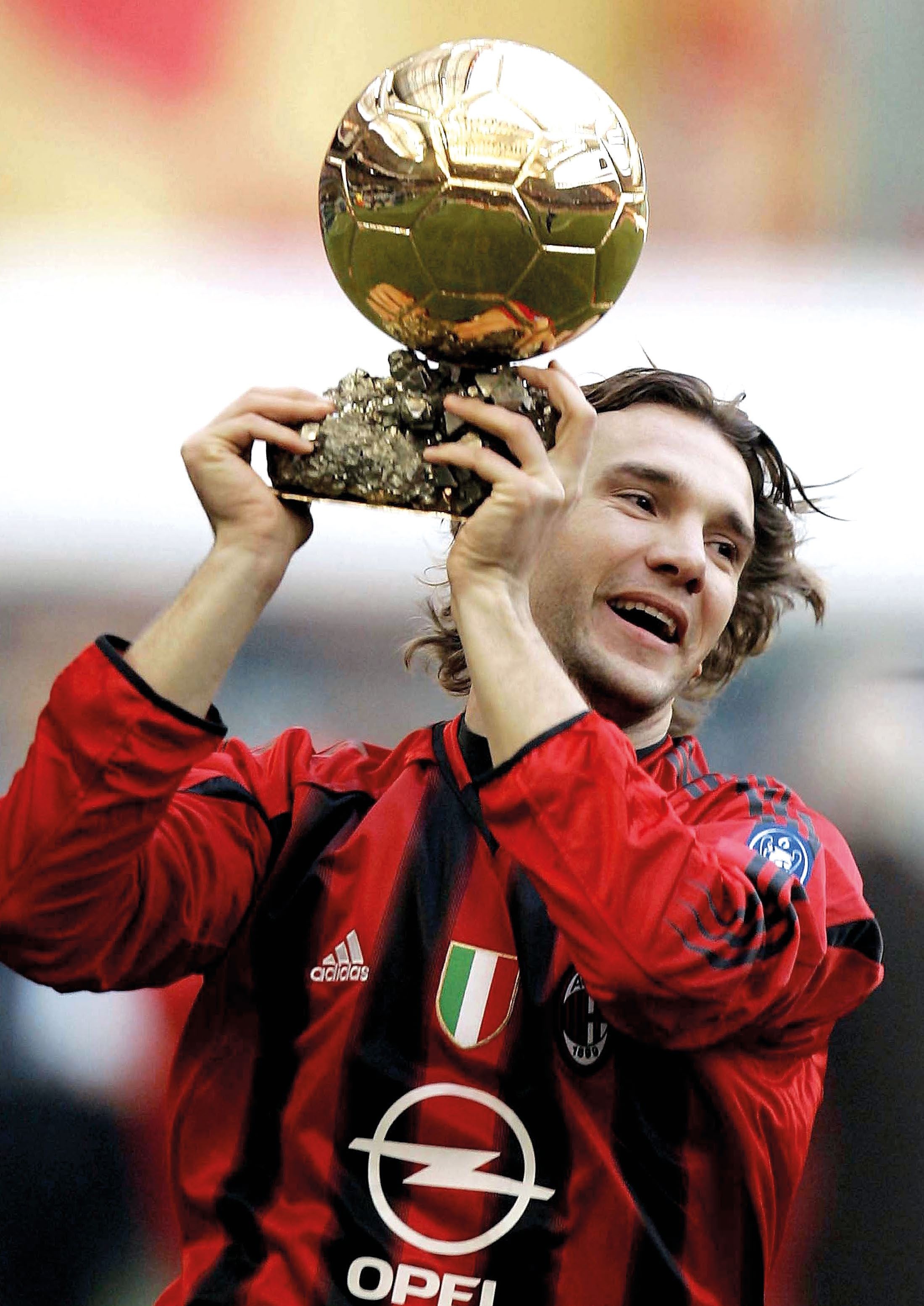 A football player in a red and black jersey joyfully holds up the Ballon d'Or trophy, celebrating a significant achievement.