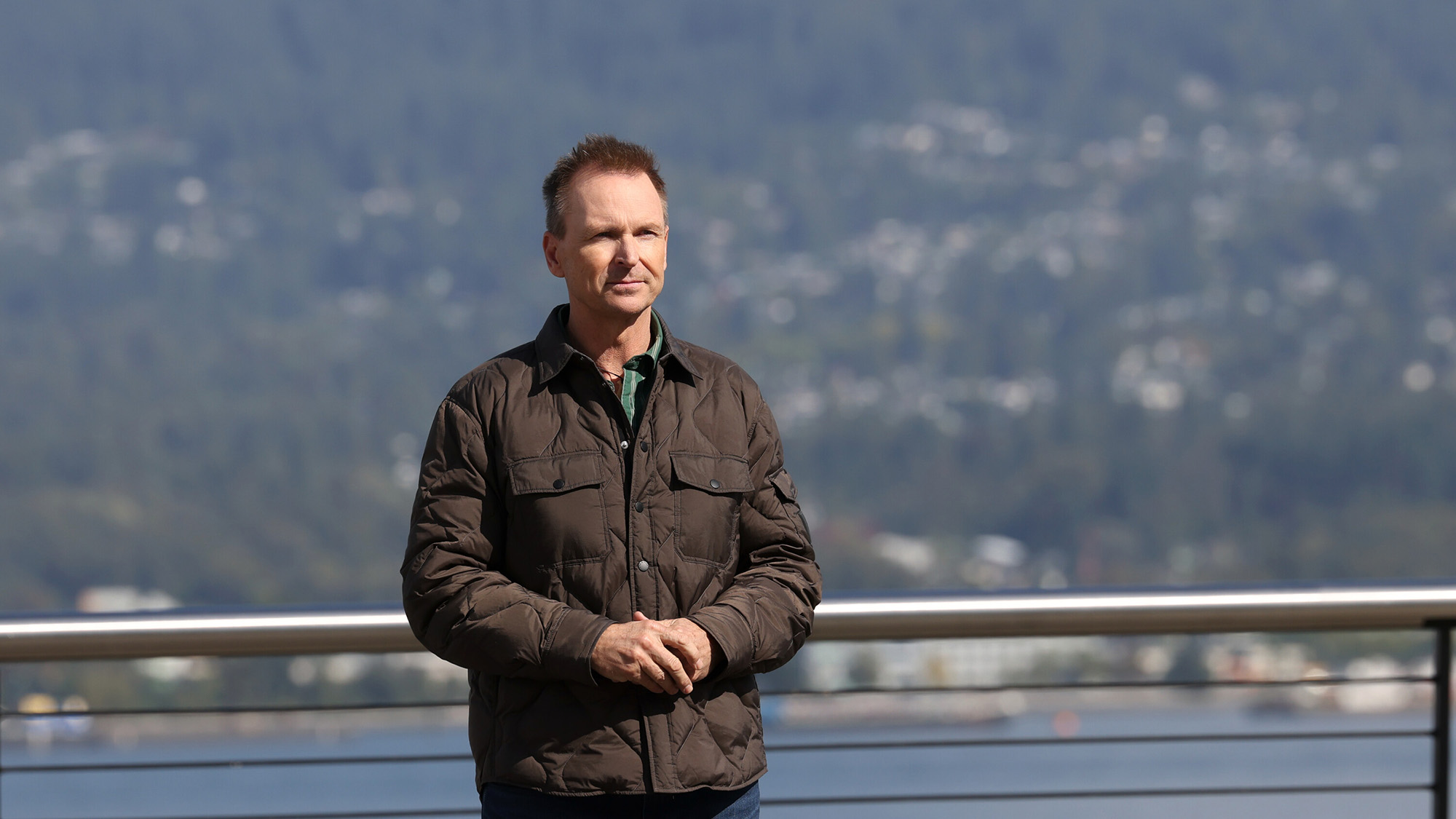 Phil stands on the finish line of an elimination leg wearing a brown jacket. Water and a mountain are in the background. 