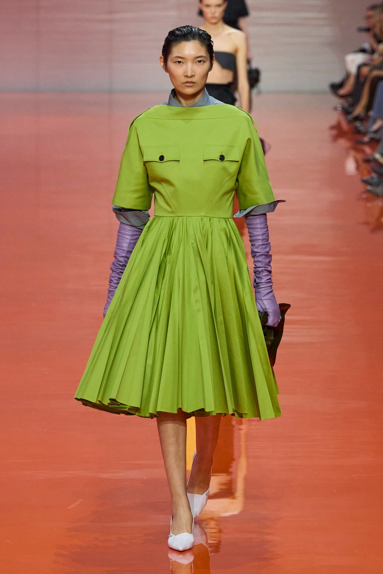 A Prada model wearing a slime green boat-neck dress with a full pleated skirt styled with a gray shirt underneath, bright purple leather opera gloves, a black clutch bag, and white pointed heels.