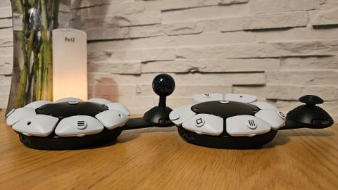 Two PlayStation Access controllers on a wooden surface with a candle, plant vase, and white brick wall in the background